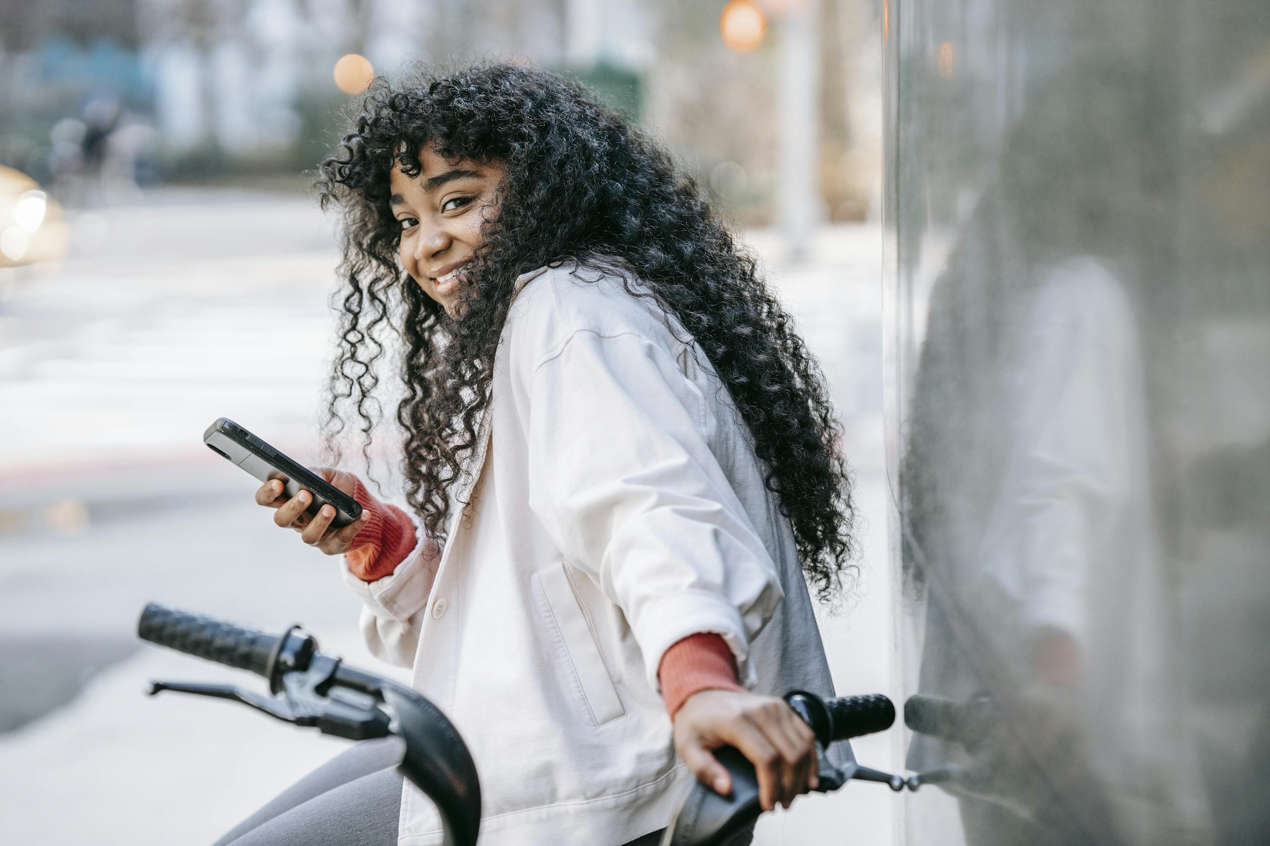 A cheerful woman with curly hair sits on a bicycle, smiling while using her smartphone outdoors.